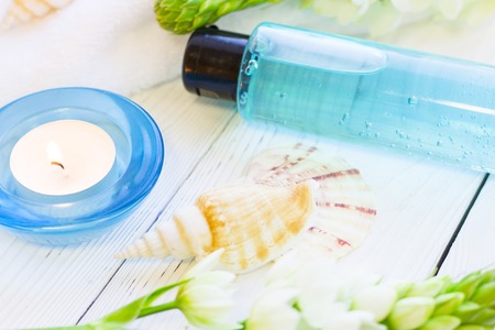 Spa items set with a blue bottle of showel gel, sea shells, flower spur,a burning candle and clean towels, white wooden background, close upの写真素材