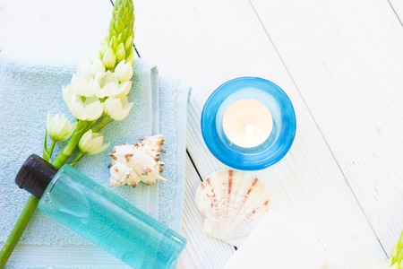 Spa items set with a blue bottle of shower gel, sea shells on a blue towel, a burning candle and a white flower spur, white wooden background, close upの写真素材