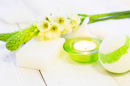 Spa and Floral concept, a spur of white flowers, a bar of soap, a fragrant bath bomb and a burning candle in a green glass holder, white wooden backgroundの写真素材
