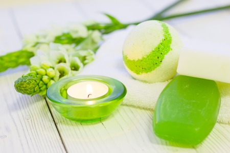 Spa and Floral concept, a spur of white flowers, a fragrant bath bomb, fruit soap and a burning candle in a green glass holder, white wooden background, close upの写真素材