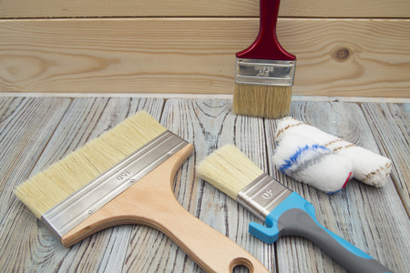 A set of paint brushes and rollers on a grey wooden table against a light wooden wall, close up