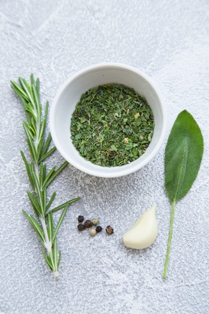 Dry herbs in a ceramic bowl, fresh herbs and garlic clove on a light stone background with copy...