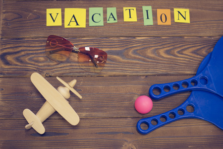 Travel, Sport Concept. Vacation inscription, blue plastic tennis rackets, a ball, toy wooden plane and sunglasses on a wooden table, top view, copy spaceの写真素材