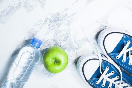 Time for diet slimming weight loss concept. Sport fitness, apple, sneakers, bottle of water and yellow weights on white stone background.の写真素材