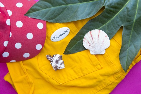 Vacation concept. Folded beach clothes and sea shells on pink background with a palm leaf