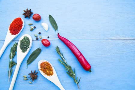 Cooking Hot Spicy Food Concept. Dry spices and herbs in wooden spoons, fresh herbs,chili peppers and cherry tomatoes on blue wooden background, top view, mock upの写真素材