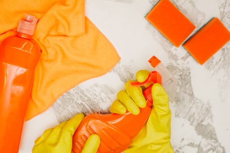 Cleaning house or office concept. Hands in yellow rubber gloves holding a spray bottle, orange cleaning supplies, white stone background, top viewの写真素材