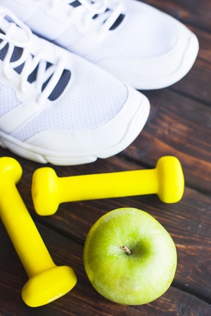 Time for diet slimming weight loss concept. Sport fitness, apple, sneakers, bottle of water and yellow weights on dark wooden background.の写真素材
