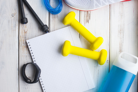 Sport fitness, apple, sneakers, bottle of water and yellow weights on white wooden background.