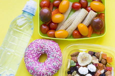 School lunch box. Donat, a bottle of water, candies, baby corns, carrot and tomatoes in green...