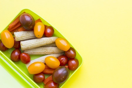 School lunch box. Candies, baby corns, carrot and tomatoes in green plastic container. Top view,...