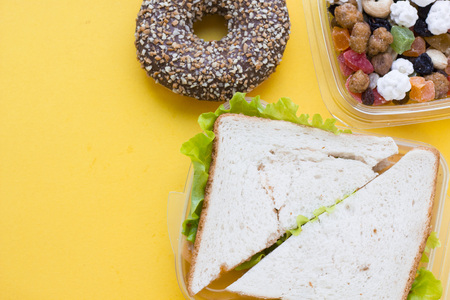 School lunch box. Sandwich, a banana, candies, baby corns, carrot and tomatoes in green plastic container. Top view, yellow backgroundの写真素材