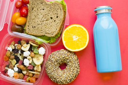 School lunch box. Bread, orange, blue bottle, baby corns, carrot and tomatoes in green plastic container. Top view, red backgroundの写真素材