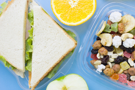 School lunch box. Bread, orange, candies, baby corns, carrot and tomatoes in green plastic container. Top view, blue backgroundの写真素材