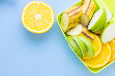 School lunch box. Bread, orange, candies, baby corns, carrot and tomatoes in green plastic container. Top view, blue backgroundの写真素材