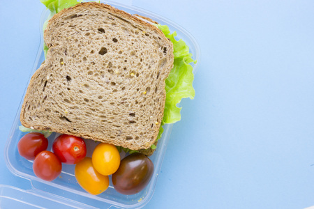 School lunch box. Bread, orange, candies, baby corns, carrot and tomatoes in green plastic container. Top view, blue backgroundの写真素材