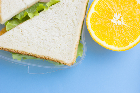 School lunch box. Bread, orange, candies, baby corns, carrot and tomatoes in green plastic container. Top view, blue backgroundの写真素材