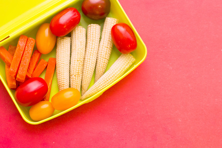 School lunch box. Bread, orange, baby corns, carrot and tomatoes in green plastic container. Top view, red backgroundの写真素材