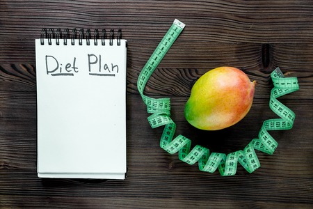 Slimming concept. Notebook for diet plan, fruit mango and measuring tape on wooden table top view mock upの写真素材