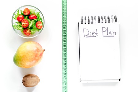 Tasty food for slimming. Notebook for diet plan, salad, fruits and measuring tape on white background top view mock upの写真素材