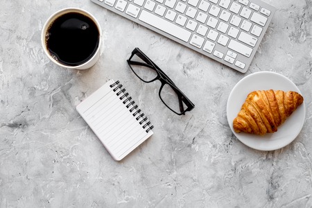 Snack for office. Coffee and croissant near keyboard, glasses and notebook on grey stone background top view copyspaceの写真素材