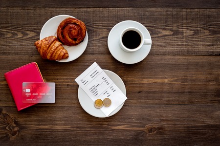 Pay bill at cafe by card. Purse, bill and bank card near coffee and croissant on dark wooden table top view copyspaceの写真素材