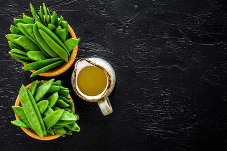 Green pea pods in bowls near jug of oil on black background top view.