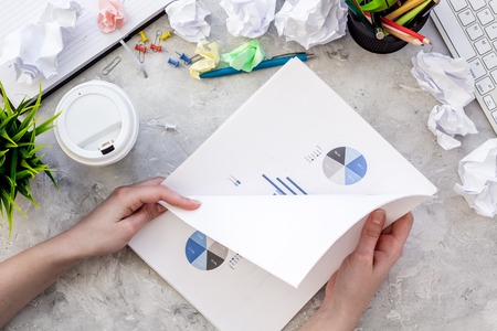 Chaos in office.Desk covered with crumpled paper and scattered stationery. Hands hold sheet with chart. Grey background top viewの写真素材