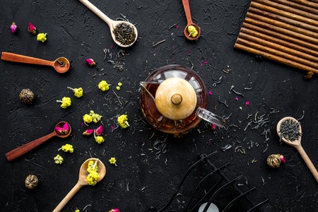 Brew the aromatic tea. Tea pot near wooden spoons with dried tea leaves, flowers and spices on black...
