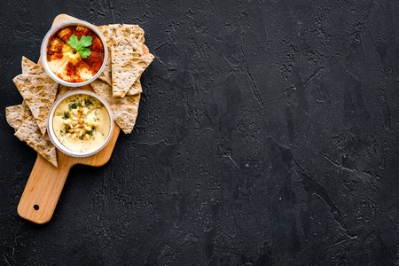 Serve hummus. Bowl with dish near pieces of crispbread on black background top view.
