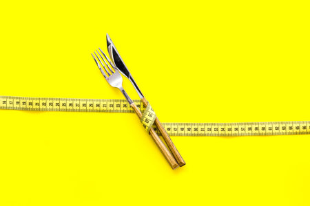 Diet, weight loss, slimming concept. Fork and knife with wound measuring tape on yellow background...