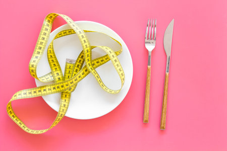 Diet for weight loss concept. Proper nutrition. Medical starvation. Empty plate with fork and knife near measuring tape on pink background top viewの写真素材