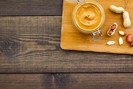 Make peanut butter. Paste in glass jar on cutting desk near peanut on dark wooden background top...