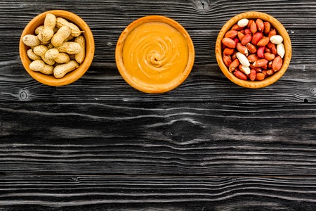 Peanut paste concept. Bowls with butter, nuts in shell, peeled nuts on black wooden background top...