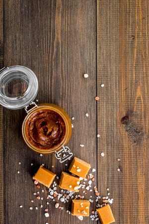 Contrast of flavors. The combination of salty and sweet. Caramel sauce in glass jar near caramel cubes on dark wooden background top view copy spaceの写真素材