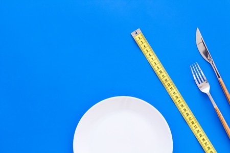 Diet for weight loss concept. Proper nutrition. Medical starvation. Empty plate with fork and knife near measuring tape on blue background top view mockupの写真素材