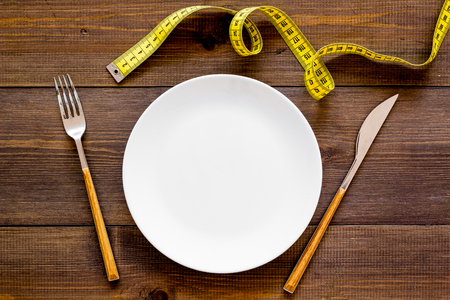 Diet for weight loss concept. Proper nutrition. Medical starvation. Empty plate with fork and knife near measuring tape on dark wooden background top view mockupの写真素材