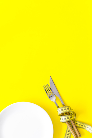 Diet for weight loss concept. Proper nutrition. Medical starvation. Empty plate with fork and knife near measuring tape on yellow background top view mockupの写真素材