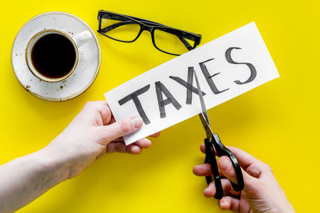 Tax reduce concept. Hands with scissors cut paper with word Taxes on yellow background with coffee and glasses top viewの写真素材