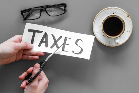 Tax reduce concept. Hands with scissors cut paper with word Taxes on grey background top viewの写真素材