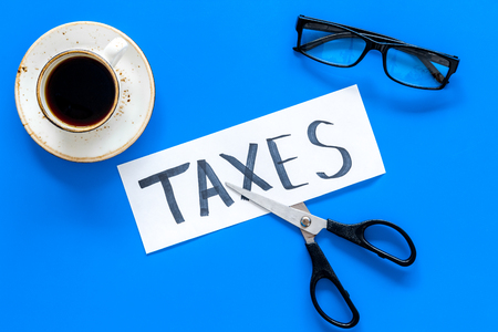 Tax reduce concept. Scissors and paper with word Taxes on blue background with coffee and glasses top viewの写真素材