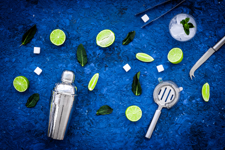 Ingredients and crockery for making mojito. Slices of lime, mint, sugar cubes, glass with ice cubes, shaker, strainer on blue background top view patternの写真素材