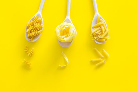 Different pasta types. Fusilli, penne, fettuccine in spoons on yellow background top view copy spaceの写真素材