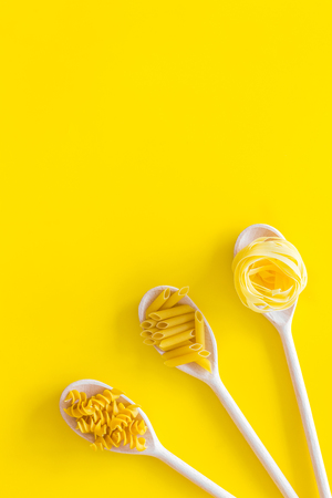 Different pasta types. Fusilli, penne, fettuccine in spoons on yellow background top view copy spaceの写真素材