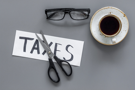 Tax reduce concept. Scissors and paper with word Taxes on grey background with coffee and glasses top viewの写真素材