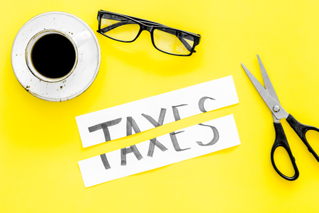 Tax reduce concept. Scissors and paper with word Taxes on yellow background with coffee and glasses top viewの写真素材