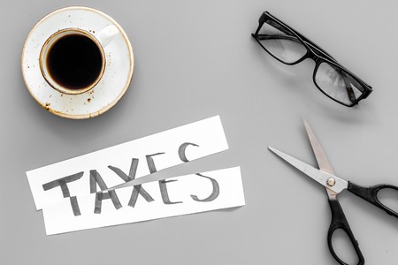 Tax reduce concept. Scissors and paper with word Taxes on grey background with coffee and glasses top viewの写真素材