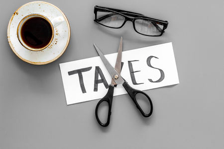 Tax reduce concept. Scissors and paper with word Taxes on grey background with coffee and glasses top viewの写真素材