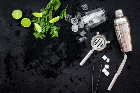 Ingredients and crockery for making mojito. Slices of lime, mint, sugar cubes, glass with ice cubes, shaker, strainer on black background top view copy spaceの写真素材