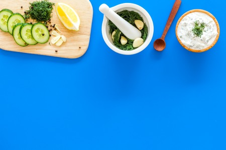 Preparing greek cucumber yogurt sauce. Bowl with yogurt near greenery, cucumber, oranges on cutting board on blue kitchen desk top view copy spaceの写真素材